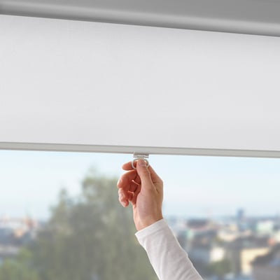 A hand adjusts a white block-out roller blind, controlling light in a room with a city view.