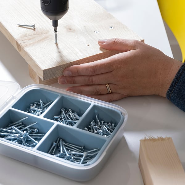 A hand uses a drill on a wooden board, placing screws from a compartmented box that holds various sizes of screws.