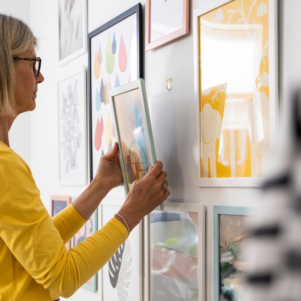 Person in yellow shirt hangs colourful picture frame on wall, using numerous tools.