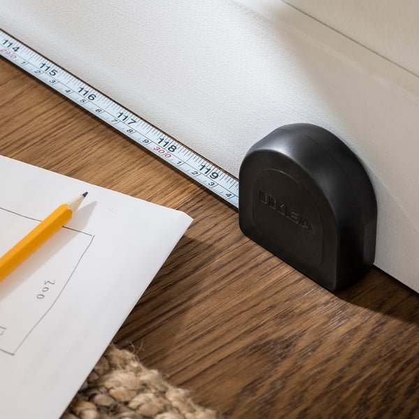 A black TRIXIG tape measure with a yellow pencil on a wooden table, showcasing its automatic return feature and locking mechanism.