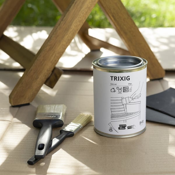 A can of clear TRIXIG wood stain sits on a wooden surface with a brush and a metal object, appearing smaller than the surrounding items.