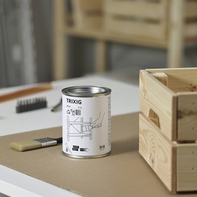 A small, round can labelled TRIXIG sits on a work surface next to a paintbrush and an unfinished wooden crate.