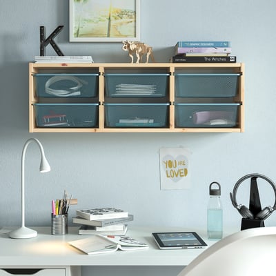 Wall-mounted TROFAST storage in light wood holds books and decor above a white desk with a lamp and tablet.