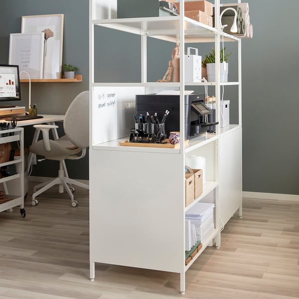 White TROTTEN double-sided cabinet in home office, storing printer, plants, boxes. Divides workspace with open and closed storage, modern design.
