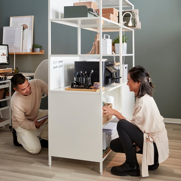 Two people organising a double-sided white cabinet with open/closed storage, serving as a room divider and workspace with printer and supplies.