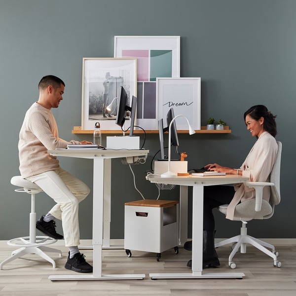 Two people using TROTTEN desks, switching between sitting and standing positions with crank handles, in a home office setup with monitors and decor.