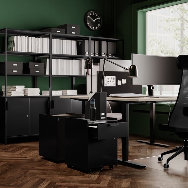 Modern home office with black TROTTEN desk, steel legs, green walls, wooden floor, and black chair.