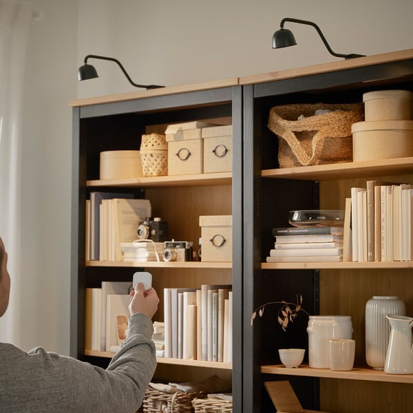 Person holding remote aiming at wall lamp above shelves. The lamp is positioned to illuminate the cabinet.