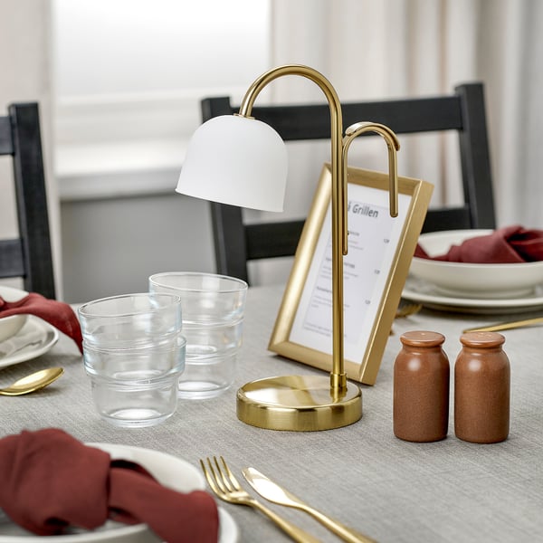A stylish, portable lamp on a table, illuminating with a warm white light, showcasing its brass base and frosted glass shade.