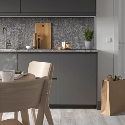 Small, sleek, gray kitchen with modern cabinets, wooden chair, and marble backsplash. A cutting board, bowls, and small plant sit on the counter.