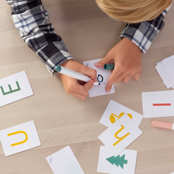 Child holding a marker, pointing at a card with letters on a wooden table, surrounded by colourful cards with letters and numbers.