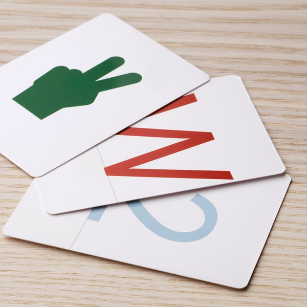 Three white educational cards lay on a wooden surface, featuring green hand symbol, red diagonal stripes, and blue letter j.