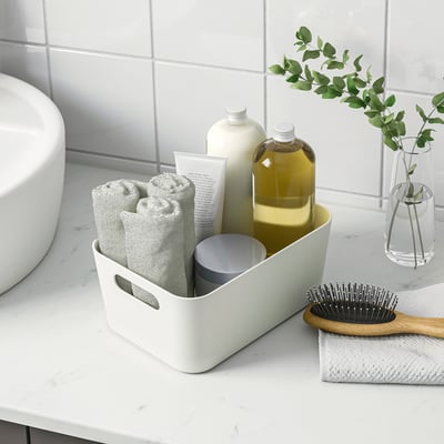 White storage box organises bathroom items like towels, bottles, and a brush. Handles make it easy to carry.
