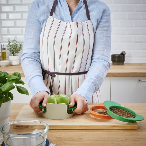 Person wearing an apron uses a green fruit cutter to slice an apple on a wooden cutting board.
