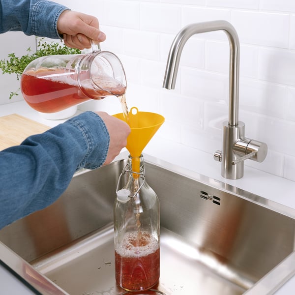 Person pours liquid from jug into glass bottle using yellow funnel in sink.