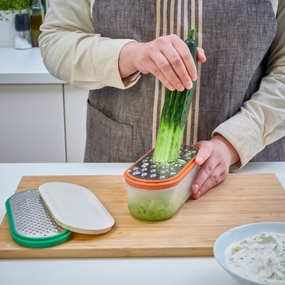 A person grates a cucumber directly into a UPPFYLLD container on a wooden cutting board.