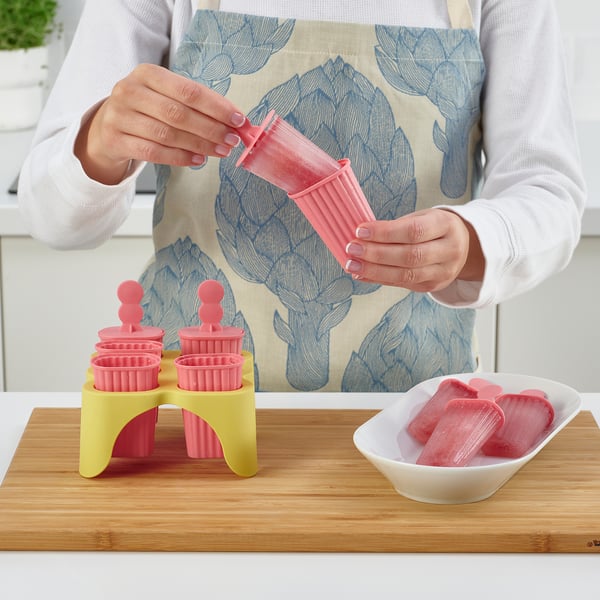 Person making popsicles using pink moulds. The person pours liquid into moulds held by a yellow stand. Completed red popsicles in a white dish nearby.