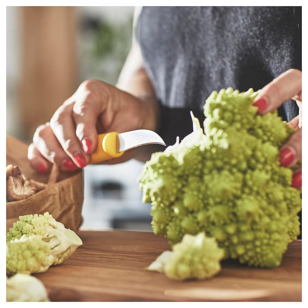 Person cutting green vegetable with small, yellow-handled knife on wooden board.