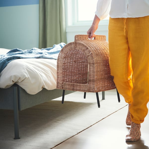 Person in yellow pants holding UTSÅDD cat house, showing its portable handle and elevated design for air circulation.