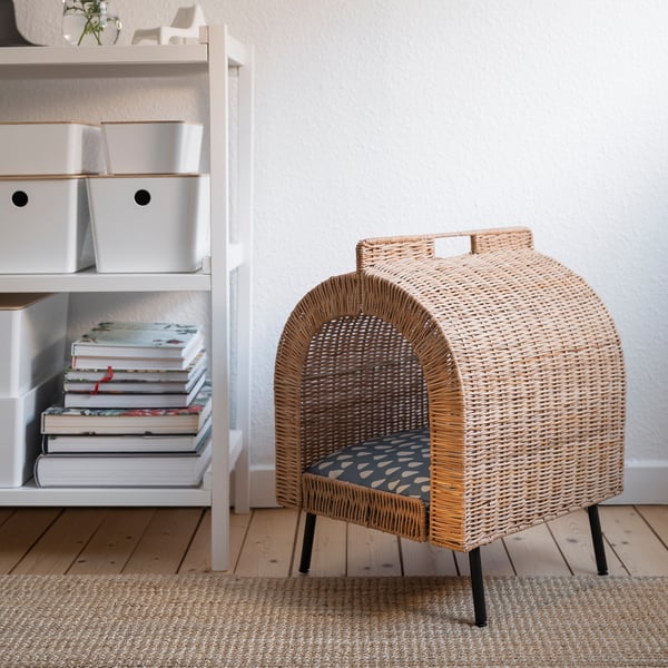 Wicker cat house on rug near white shelves with books and storage bins.