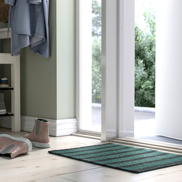 Bright hallway with large VÄGSKYLT door mat, blue-green stripes, placed under glass doors, smaller pink boots beside.