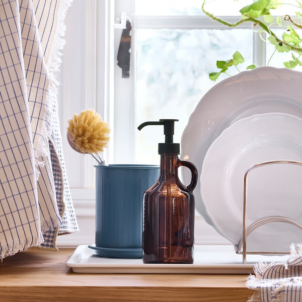 Window side, wooden counter holds a dish brush in blue cup, soap dispenser, and stacked plates on stand. Curtains hang beside window.