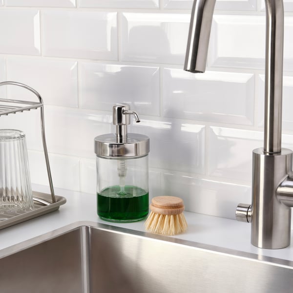 Sink with soap dispenser and scrub brush, stainless steel tap, white tiled backsplash.