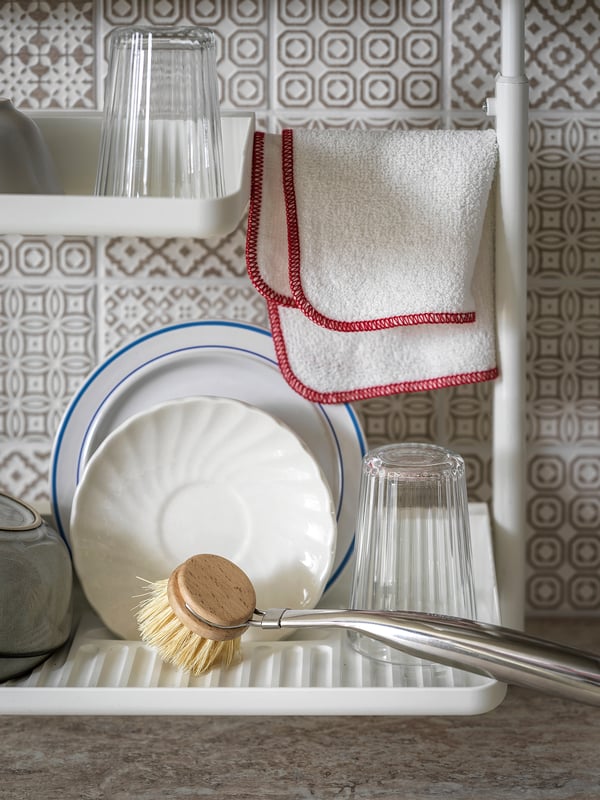 Kitchen scene with dish rack holding white dishes, glass, and VÄLVÅRDAD red bristle brush, against patterned tile wall.