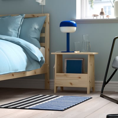 Blue and black striped rug VÄNDPLATS beside wooden bed, smaller than bed, soft low pile, rubber backing for grip.