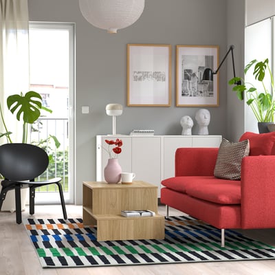 Modern living room with red sofa, black chair, striped rug, art, plants.