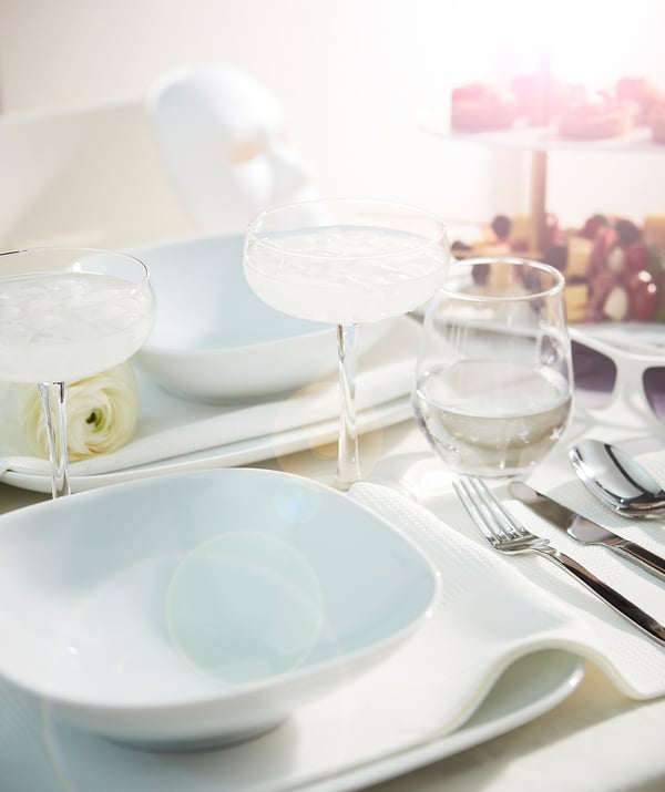 Elegant table setting with white VÄRDERA square porcelain plates, clear glass cups, silverware, and napkins. Simple, sophisticated design.