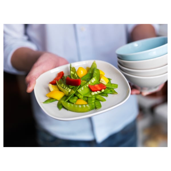 A person holds a white square side plate with green and red vegetables. Stacked plates and bowls are in the background.