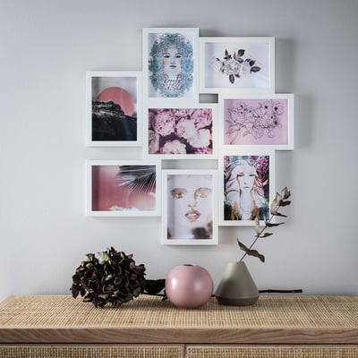 This image a wall display of seven VÄXBO frames in white, housing various artistic pictures. Below, a wooden table holds two vases and dried plants.