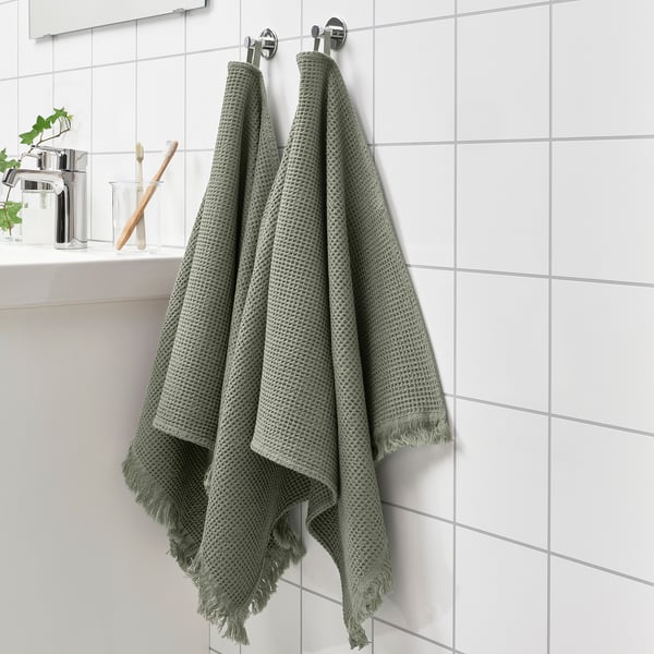 Two green VALLASÅN towels hang on wall hooks, longer than a toothbrush on nearby shelf, with light green decor.
