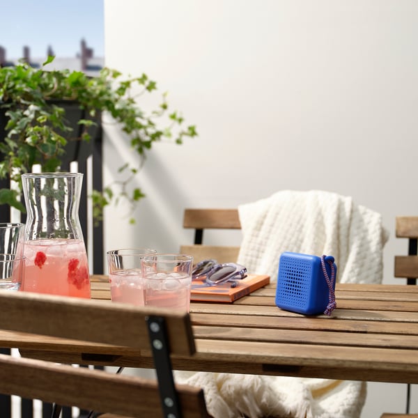 Patio table with wooden chairs, VAPPEBY speaker, drinks, and plant.