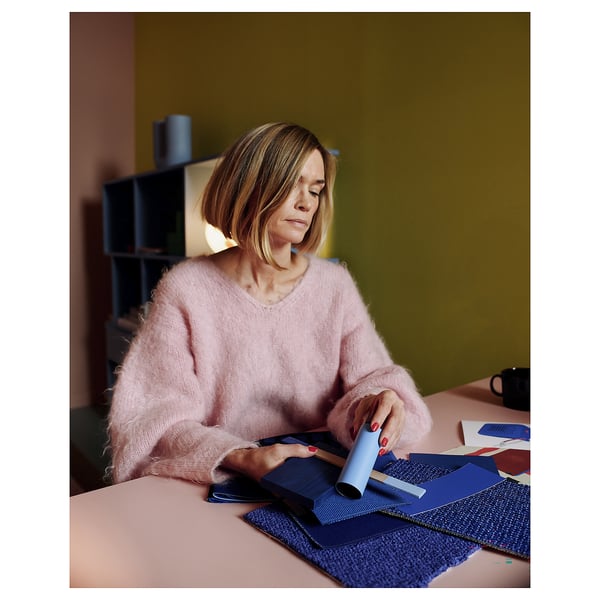 A person in a pink sweater listens to a portable blue speaker, sitting at a pink desk surrounded by fabric samples.