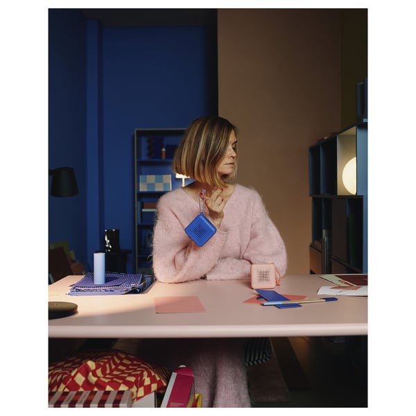 Person in pink sweater holds blue VAPPEBY speaker, sitting at a desk with colourful items in a cosy, well-lit room.