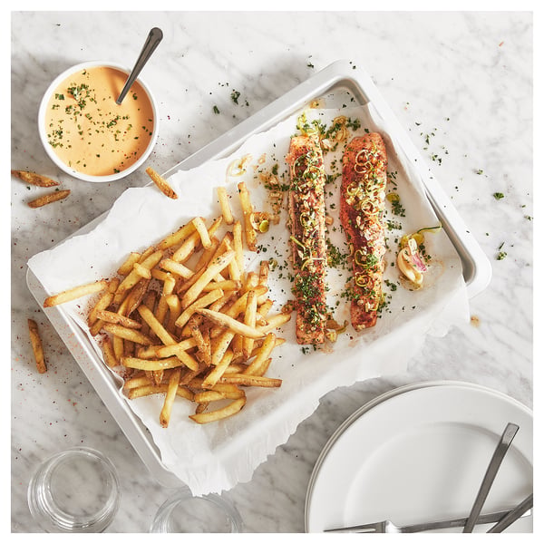 A white baking tray with golden chips and seasoned fish, a side dip, on marble surface.