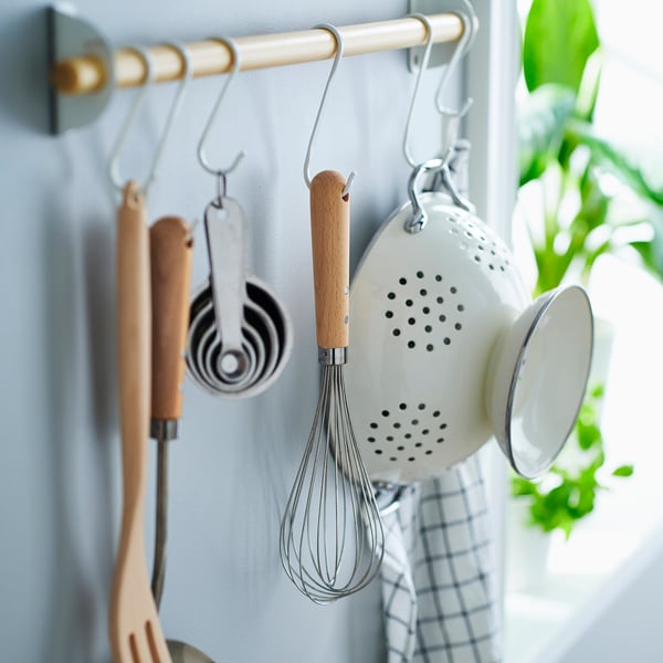 Kitchen rail with hooks holding utensils, whisk, measuring cups, and a colander. Plant on right.