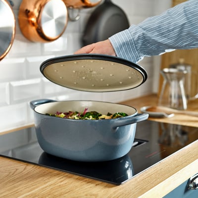 Person placing lid on large VARDAGEN casserole with veggies on modern stove.