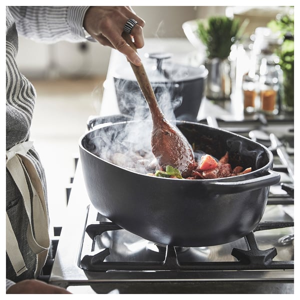 Person stirring food in a black VARDAGEN cast iron pan on a stove. Steam rises from the pan, and the cook wears an apron.