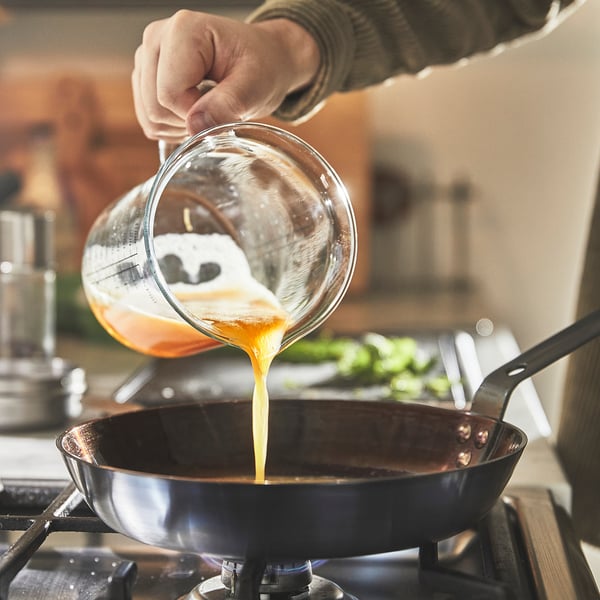 Person pours liquid from glass measuring cup into skillet on stove.