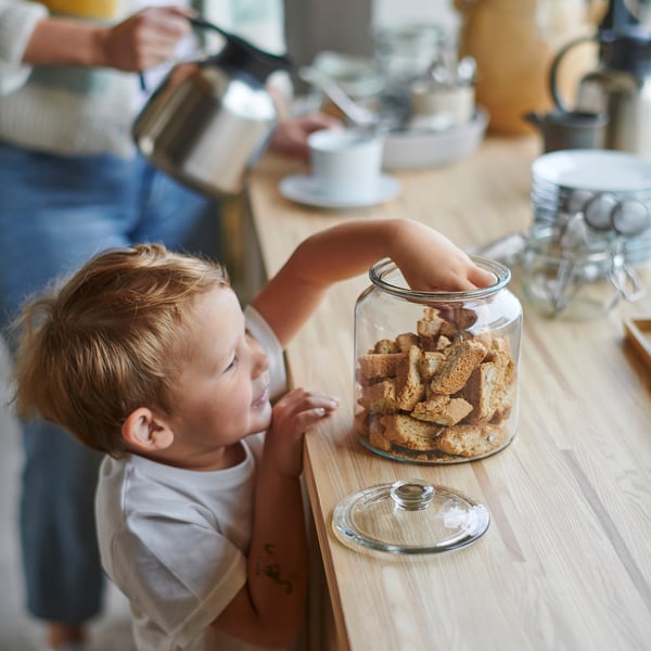 Young child reaches for cookies inside a clear glass jar on a wooden table next to teacups.