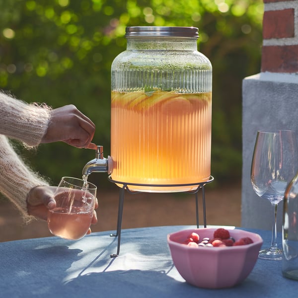 Person pours orange drink from VARDAGEN dispenser onto glass. Outdoors, table has strawberries and two glasses.