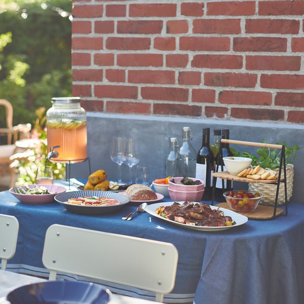 Table set outdoors: brick wall, blue cloth, chairs, dishes, drinks, glass dispenser.