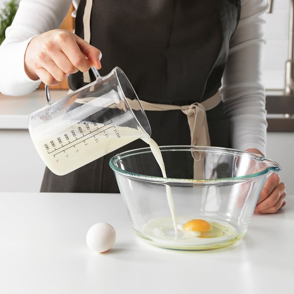Person pours milk from a measuring jug into a bowl with an egg. The jug has markings for measuring liquid.