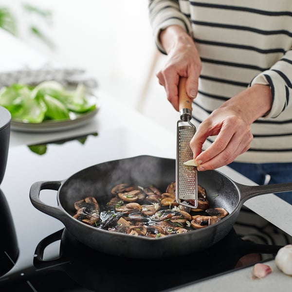 A person grates cheese over a pan of mushrooms using a wooden-handled grater. The pan is on a stove, and theres a plate of greens nearby.