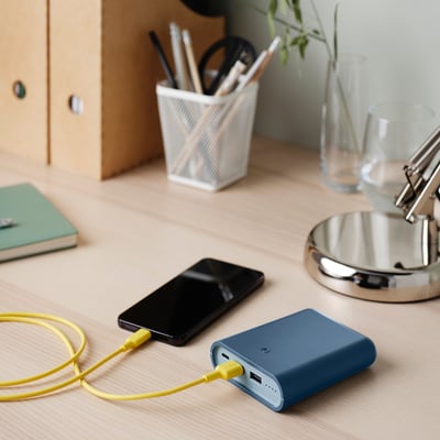 Blue power bank on desk, charging phone with yellow cable. Smaller than cup, next to pen, green notebook, and lamp.