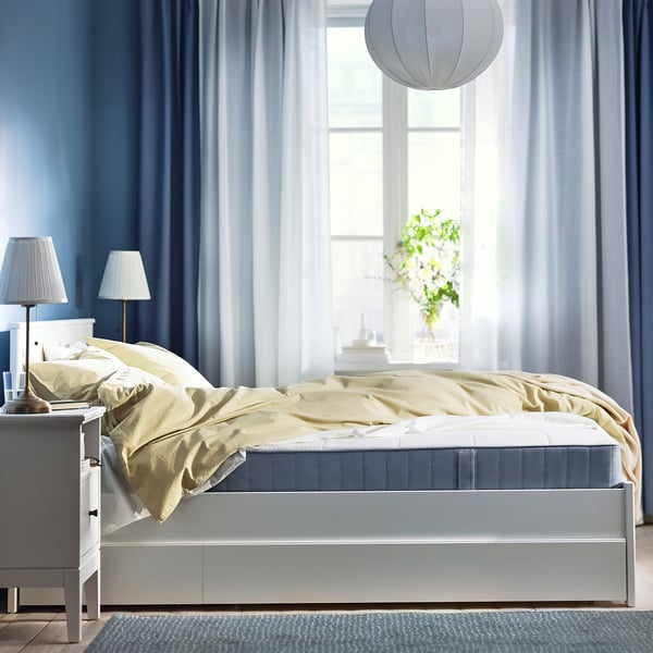 Blue bedroom with large window, modern bed, yellow blanket, two nightstands, and hanging light.