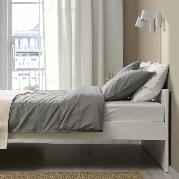 Modern bed frame with grey bedding, near a large window, includes storage, and has slatted design for airflow.